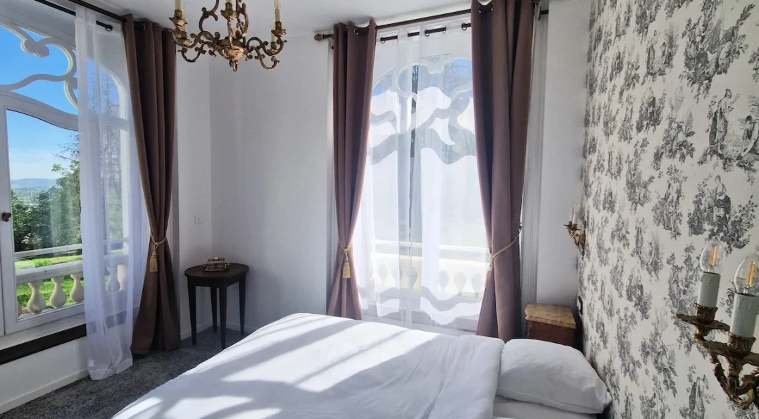Un exemple de notre chambre double château : une décoration délicate et une magnifique vue sur le parc arboré. Lit double et 2 fenêtres en angle, tapisserie motif ancien, luminaires en fer forgé patiné